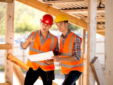Two men dressed in shirts, orange work vests and helmets explore construction documentation on the building site near the wooden building constructions